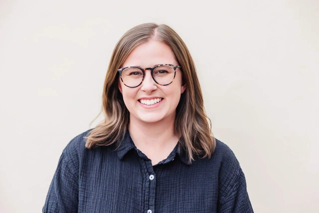 a woman smiling, wearing glasses and a dark blue shirt, with light brown hair, standing against a plain light-colored background