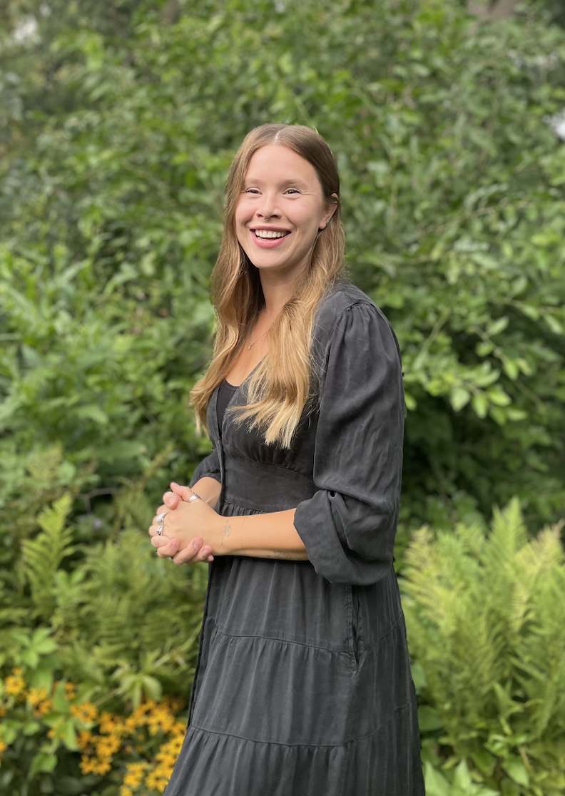 A young woman with long, light brown hair smiling and standing outdoors in front of green foliage, wearing a black dress.