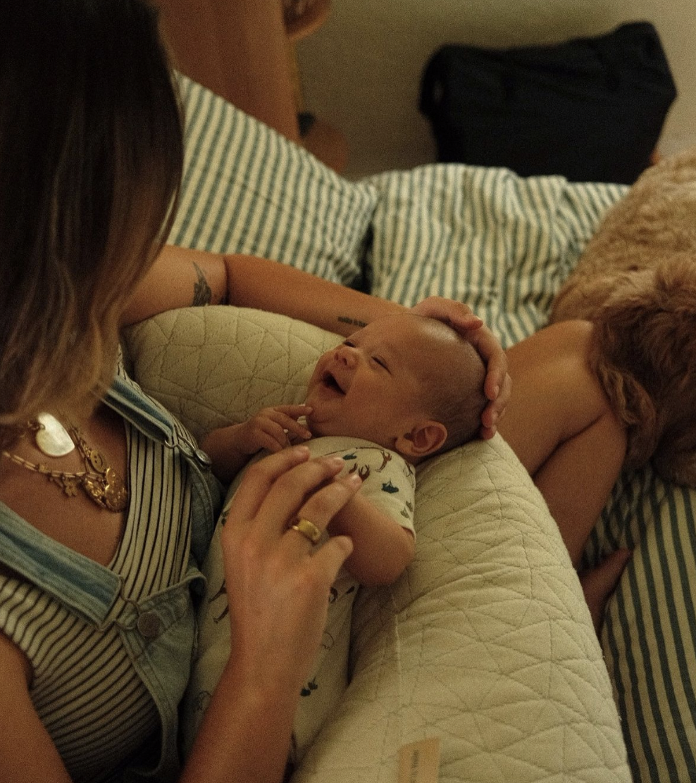 A woman lying on a bed holding a smiling baby. The woman has shoulder-length hair, wears a striped shirt, and gold jewelry. The baby is lying on the woman's lap, wearing a onesie with a pattern of animals, and appears to be happy. A dog is lying nearby on the bed, and a black bag is in the background.