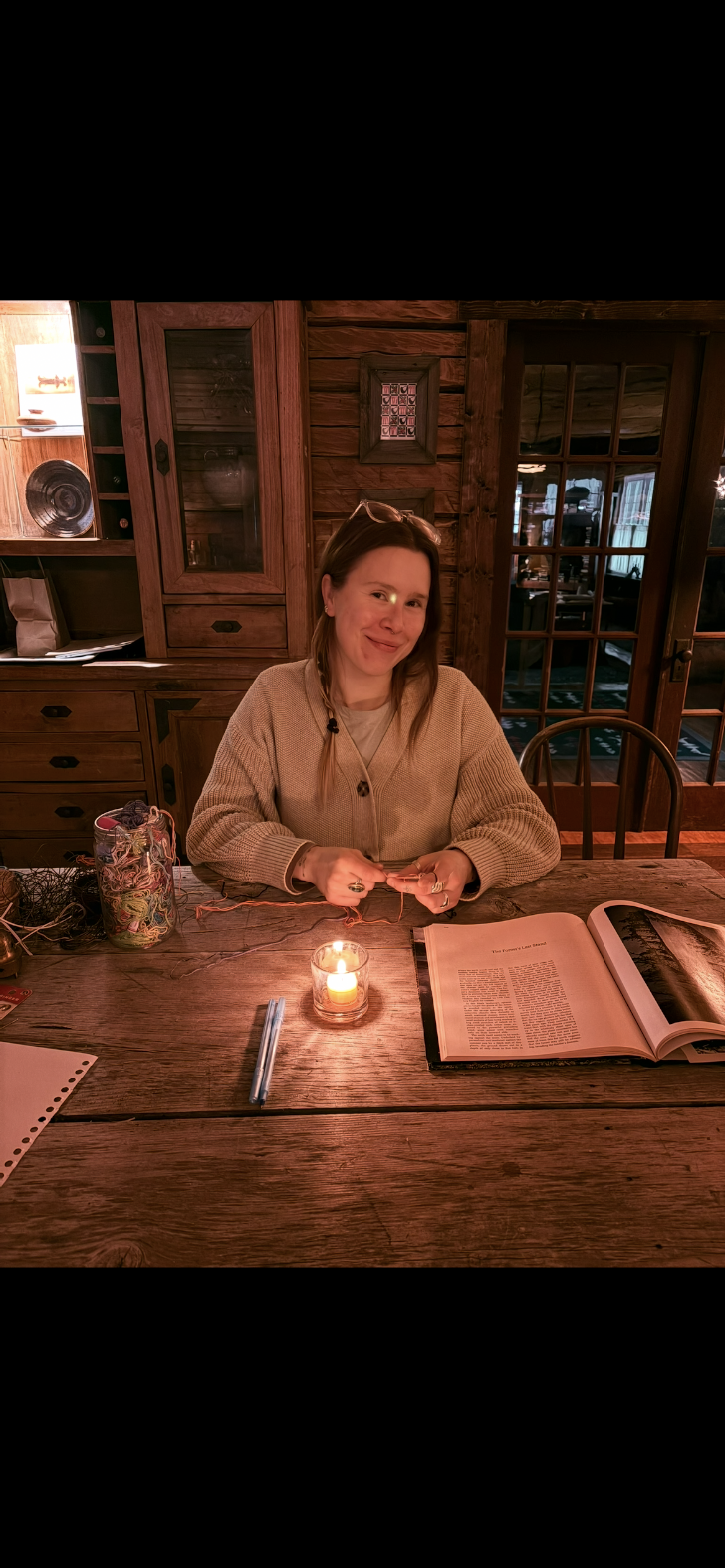 A woman sitting at a wooden table in a cozy, rustic room with wood-paneled walls, smiling at the camera. The table has a lit candle, an open book, and some yarn and supplies.