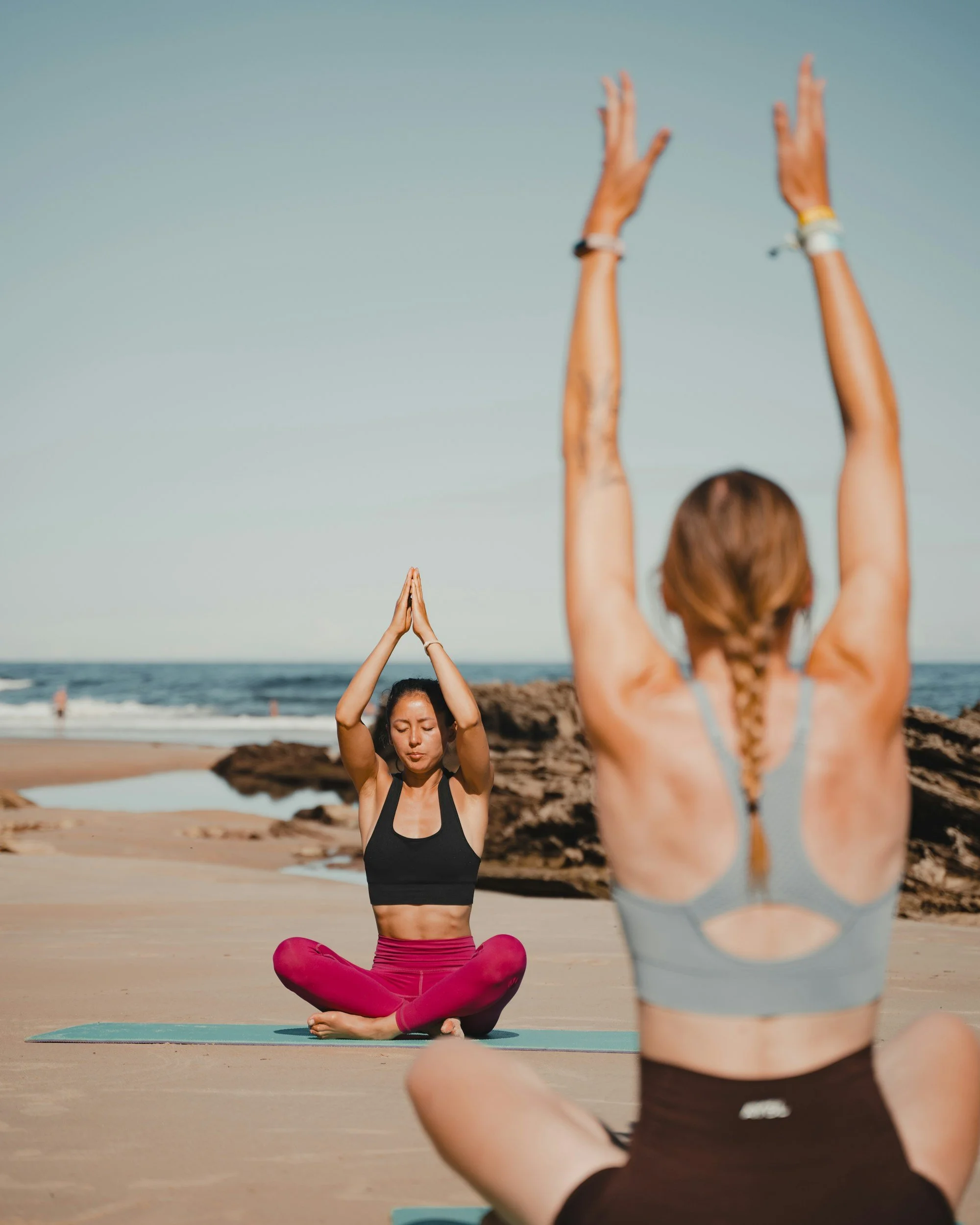 Two women practicing yoga on a beach, with the focus on the woman in the foreground who is in a seated meditation pose with her hands in prayer. The other woman is in the background, sitting cross-legged and raising her arms with palms pressed together.