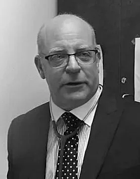 A black and white photo of an older man wearing glasses, a suit, striped shirt, and polka dot tie, standing indoors.