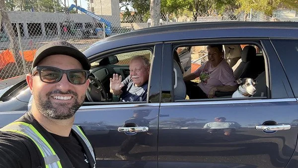 A man taking a selfie outside a moving dark-colored SUV with three passengers inside, including an elderly man waving, a woman, and a dog in a crate in the back seat.