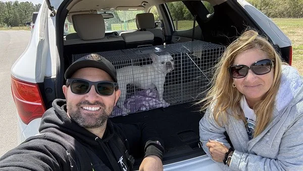 A man and woman taking a selfie in front of an open car trunk with a dog in a cage inside.