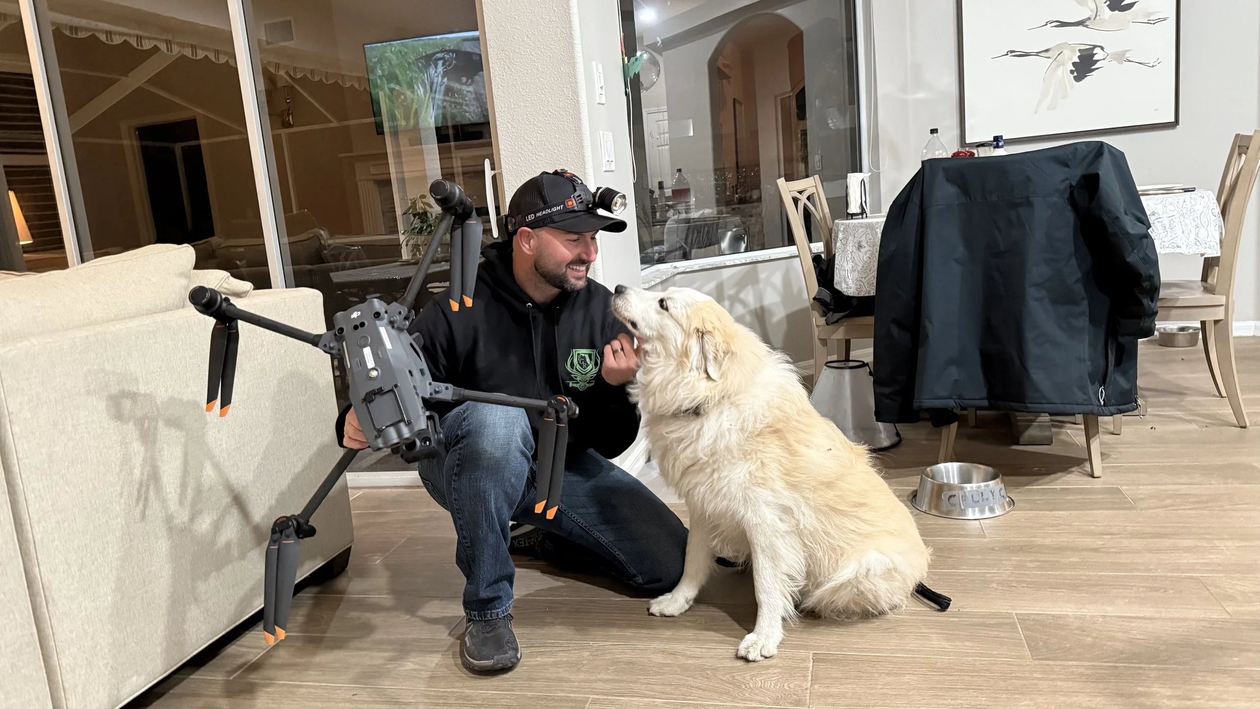 A man with a headlamp and camera equipment kneels on the floor, smiling and petting a Golden Retriever inside a home. The dog sits facing the man near its food and water bowls.