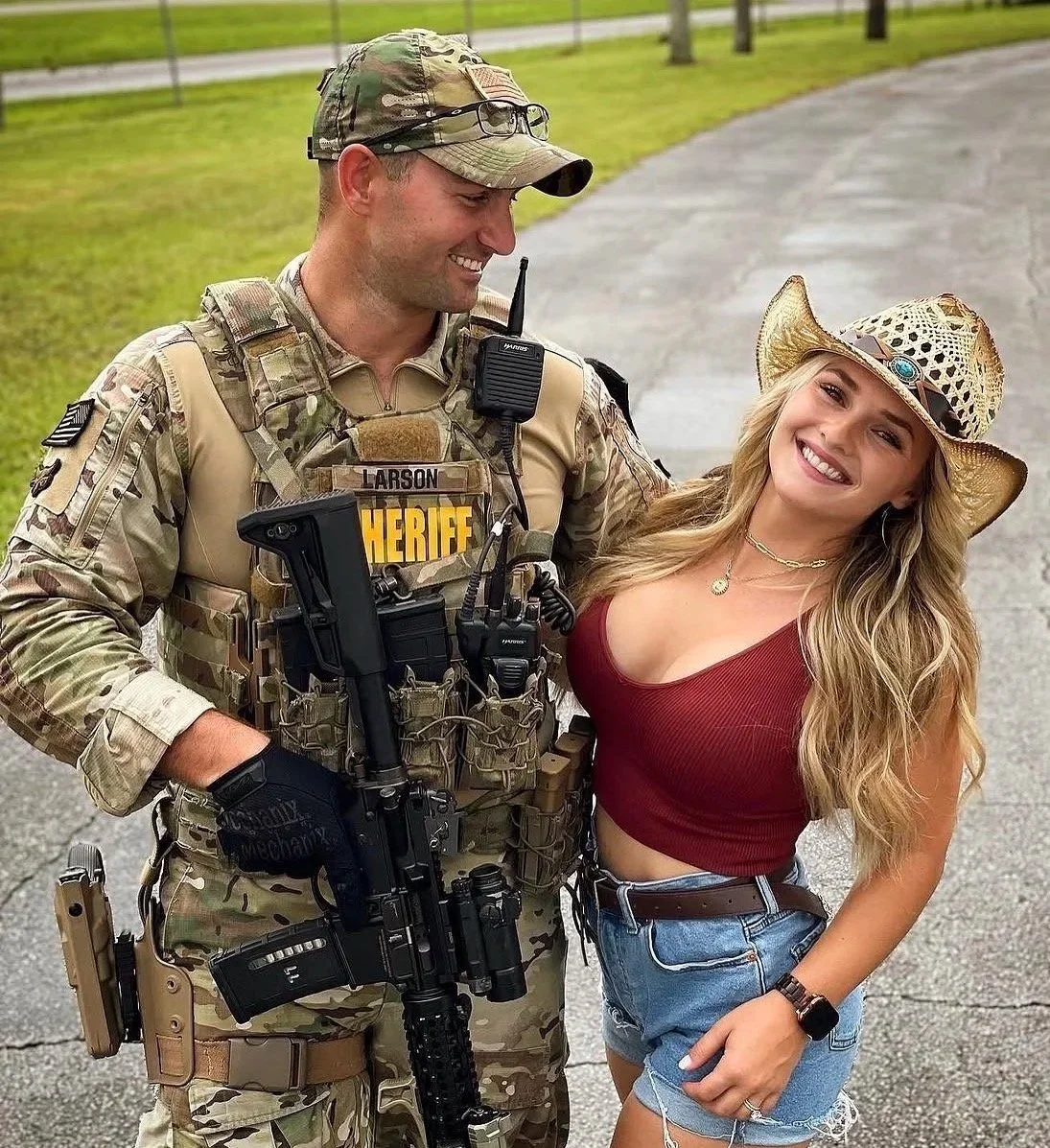 A male sheriff in camouflage uniform and a female woman in a red tank top and denim shorts smiling together outdoors, with the sheriff holding a rifle and wearing tactical gear, both looking happy.