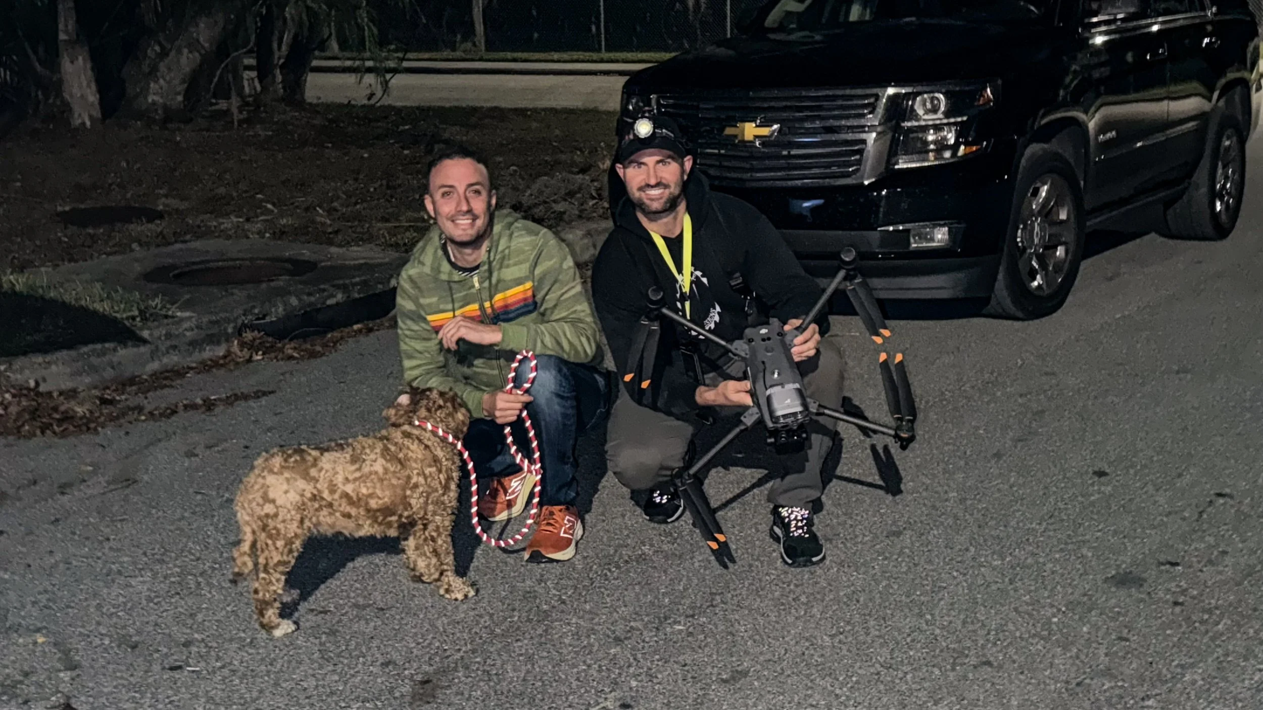 Two men kneel on a dark street at night next to a black Chevrolet SUV. One man holds a drone controller and is dressed in a black hoodie and baseball cap. The other man wears a green jacket with rainbow stripes and is holding a dog leash with a brown dog. They are smiling at the camera.