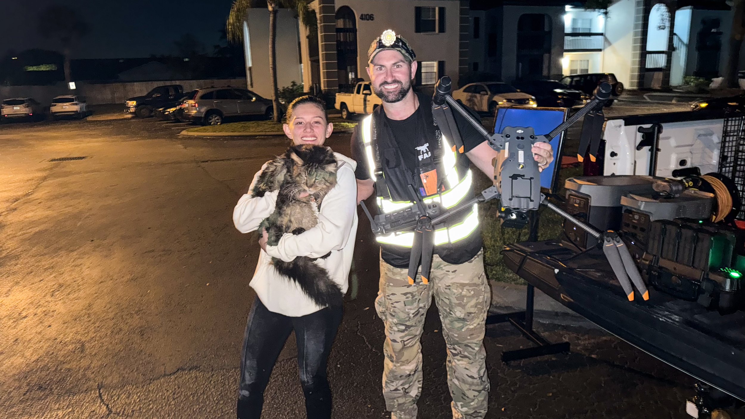 A young girl holding a large, fluffy cat and a man dressed in military camouflage with a headlamp and a drone, standing outside at night near a parking lot and an apartment complex.