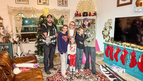 Family gathered in living room for Christmas, standing near decorated tree and fireplace with stockings.