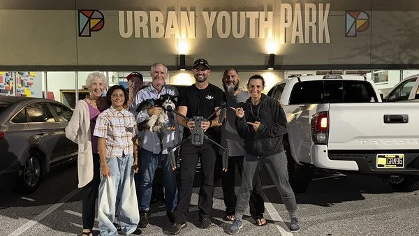 Group of seven diverse people, including children and adults, standing in front of a white pickup truck at Urban Youth Park, with a sign and parked cars in the background.