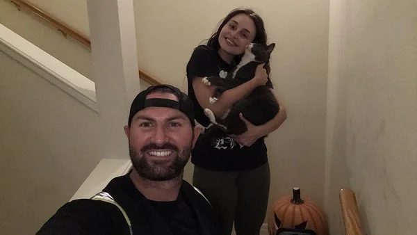 A man taking a selfie with a woman holding a black and white cat, and a pumpkin on the floor beside them.