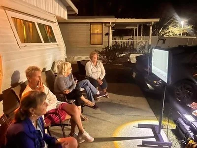 Five people sitting outside in front of a house watching a large television set on a stand, at night.