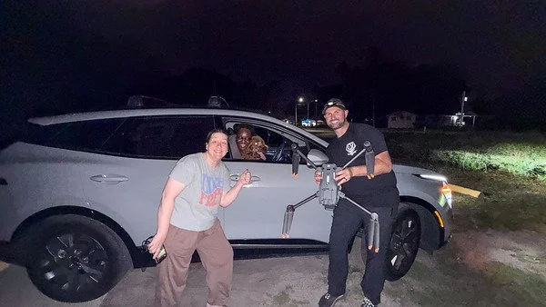 Three people, two women and a man, posing in front of a silver car at night outdoors. The woman on the left is smiling and giving a thumbs-up, the person inside the car is smiling and partially visible, and the man on the right is smiling and holding