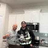Two men standing in a kitchen, smiling, holding glasses with drinks.