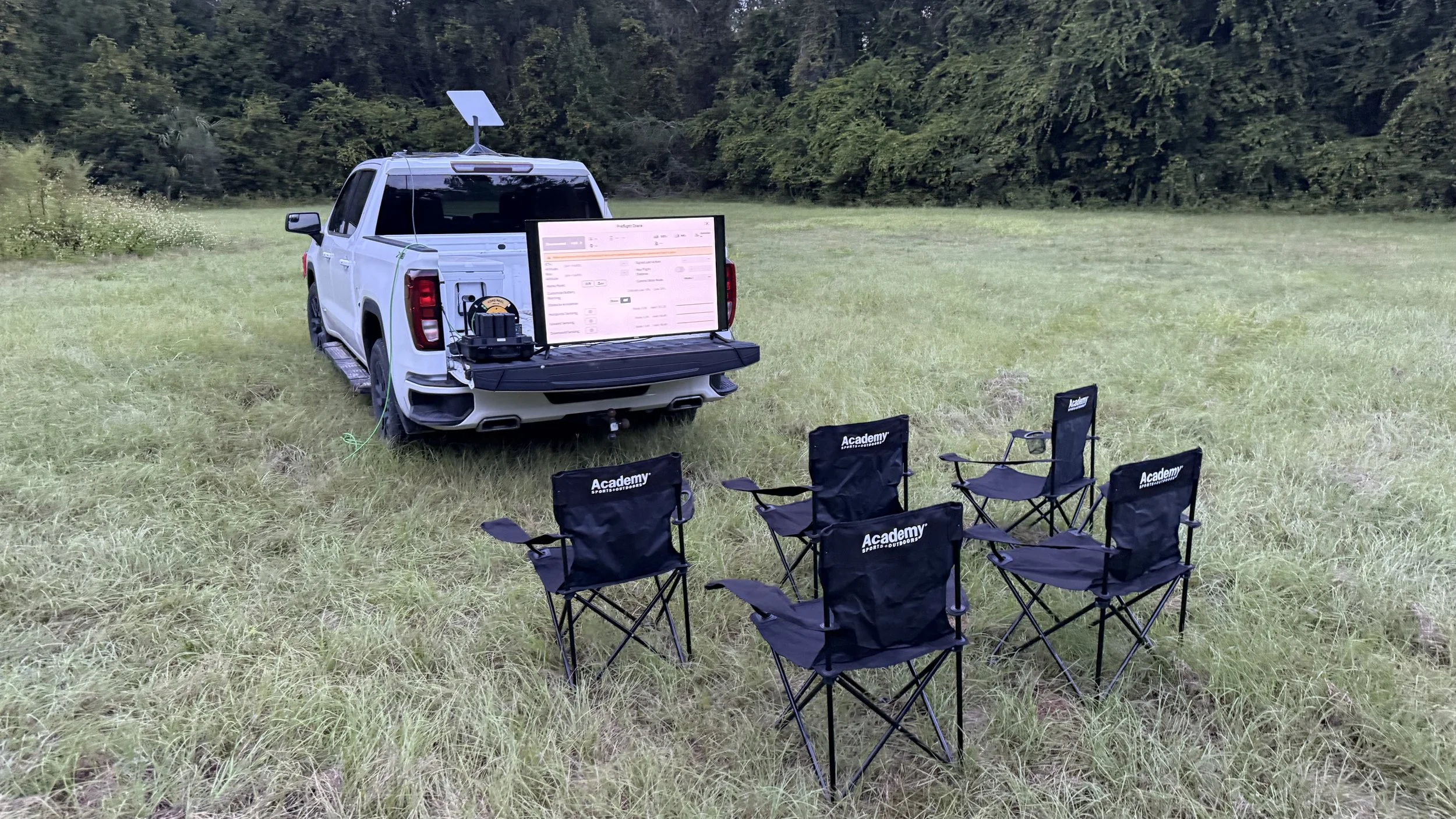 A white pickup truck parked on a grassy field with chairs and a large screen set up for outdoor activity, surrounded by trees.