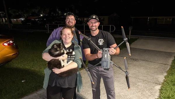 Three people standing outdoors at night, with a woman holding a black and white cat, and a man holding a large drone with four propellers.