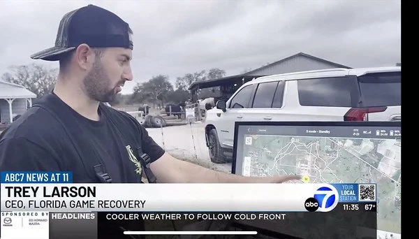 A young man with a beard wearing a black cap backward and a black t-shirt looks at a map on a tablet outdoors next to parked cars, with a cloudy sky overhead.