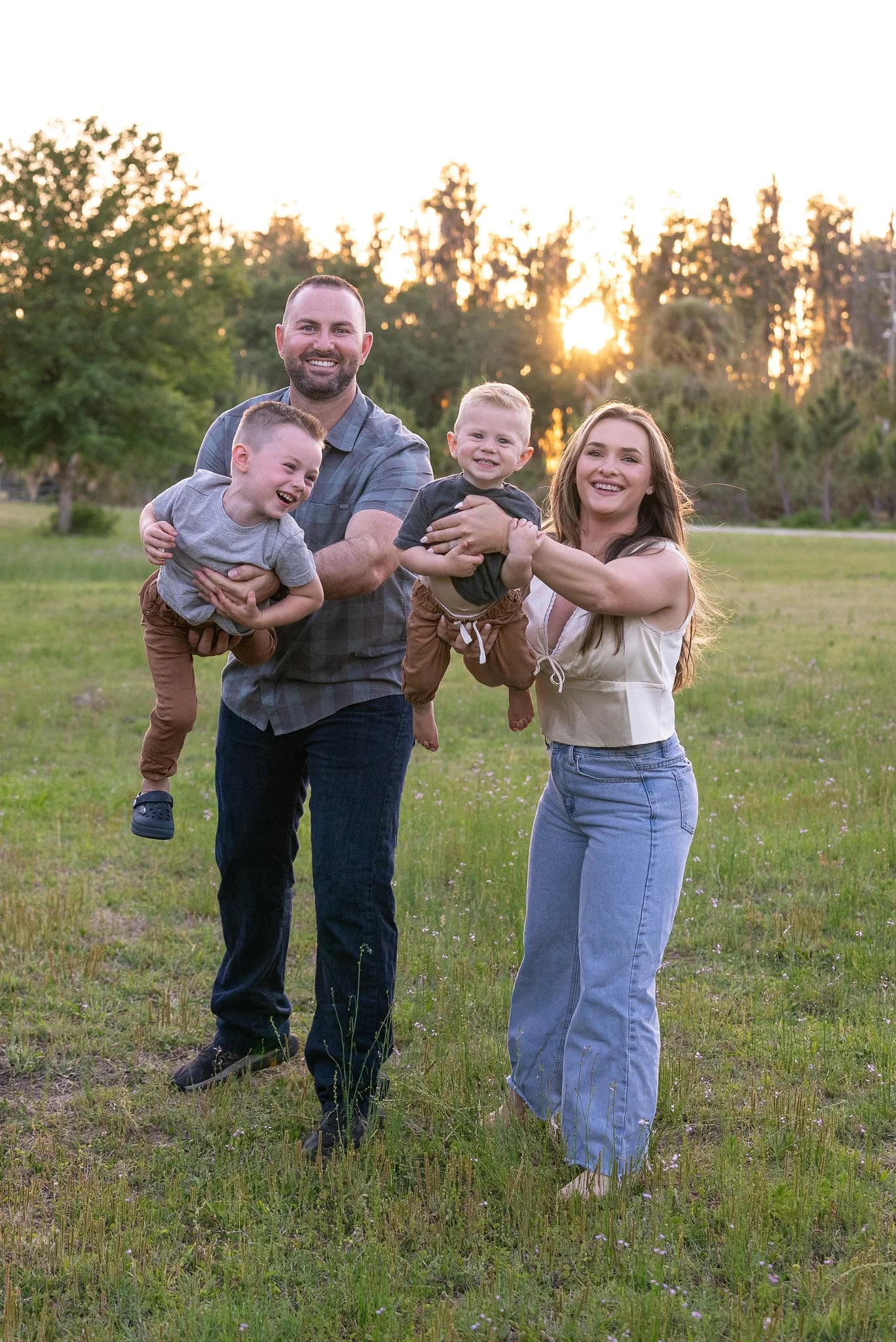 A happy family of four outdoors during sunset, with two children being held by their parents in a grassy park.