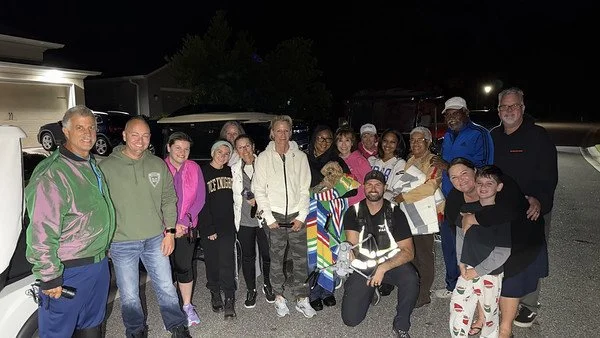 Group of people gathered outdoors at night, including children, adults, and a small dog, in a parking lot.