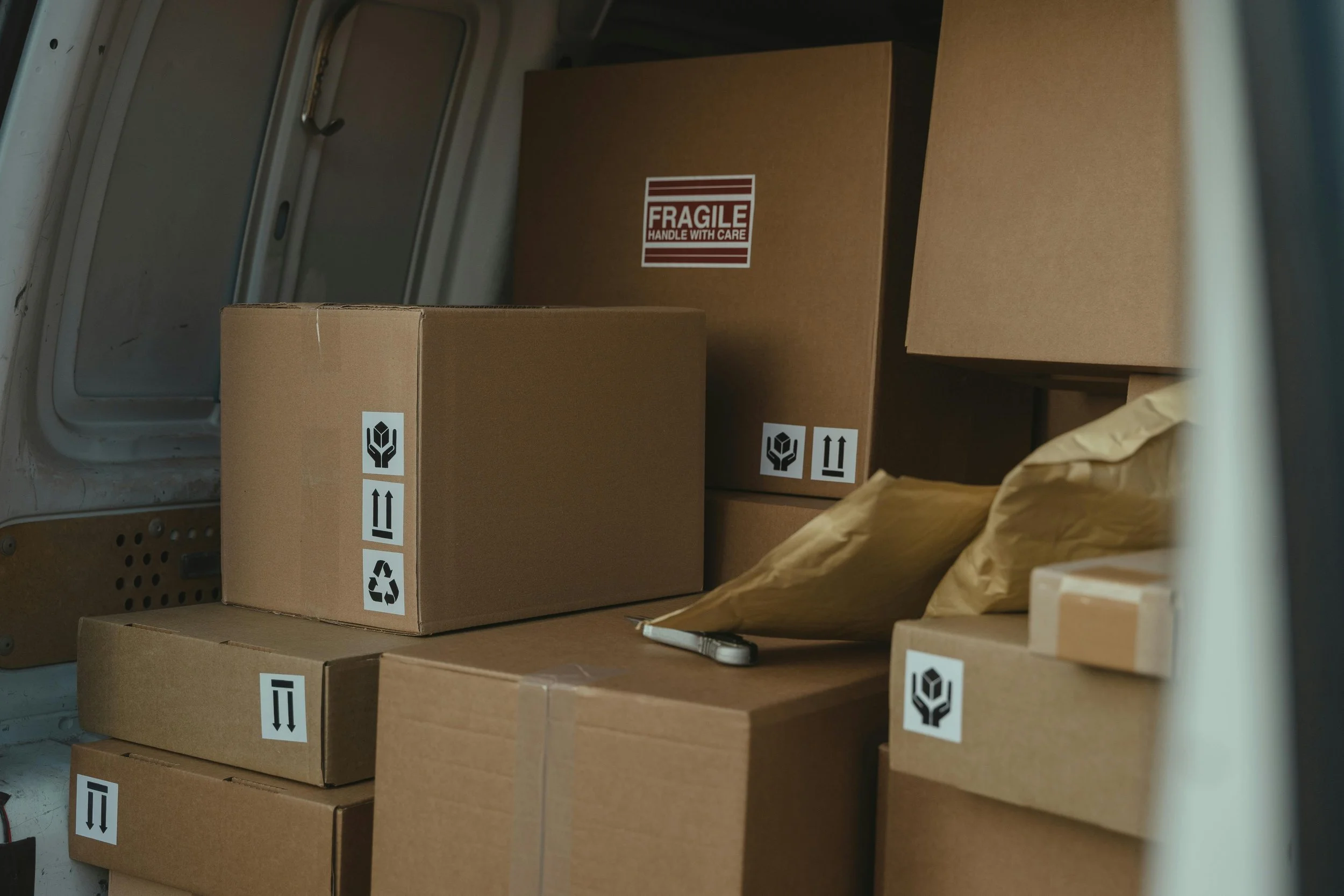 Inside a storage area with several stacked brown cardboard boxes, some labeled with fragile stickers and handling instructions, surrounded by brown paper wrapping.