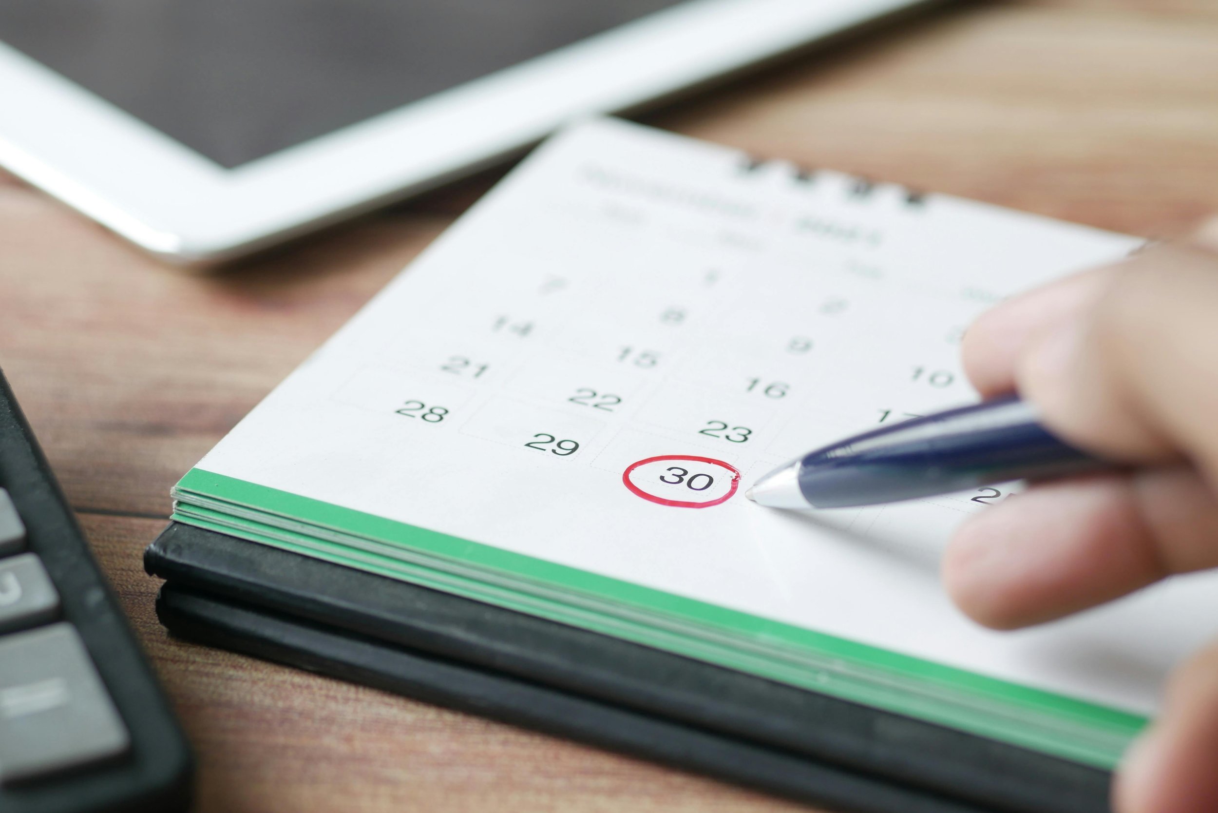 A person filling out a calendar marked with a red circle around the date 30, with a black pen, on a wooden desk near a laptop and a keyboard.