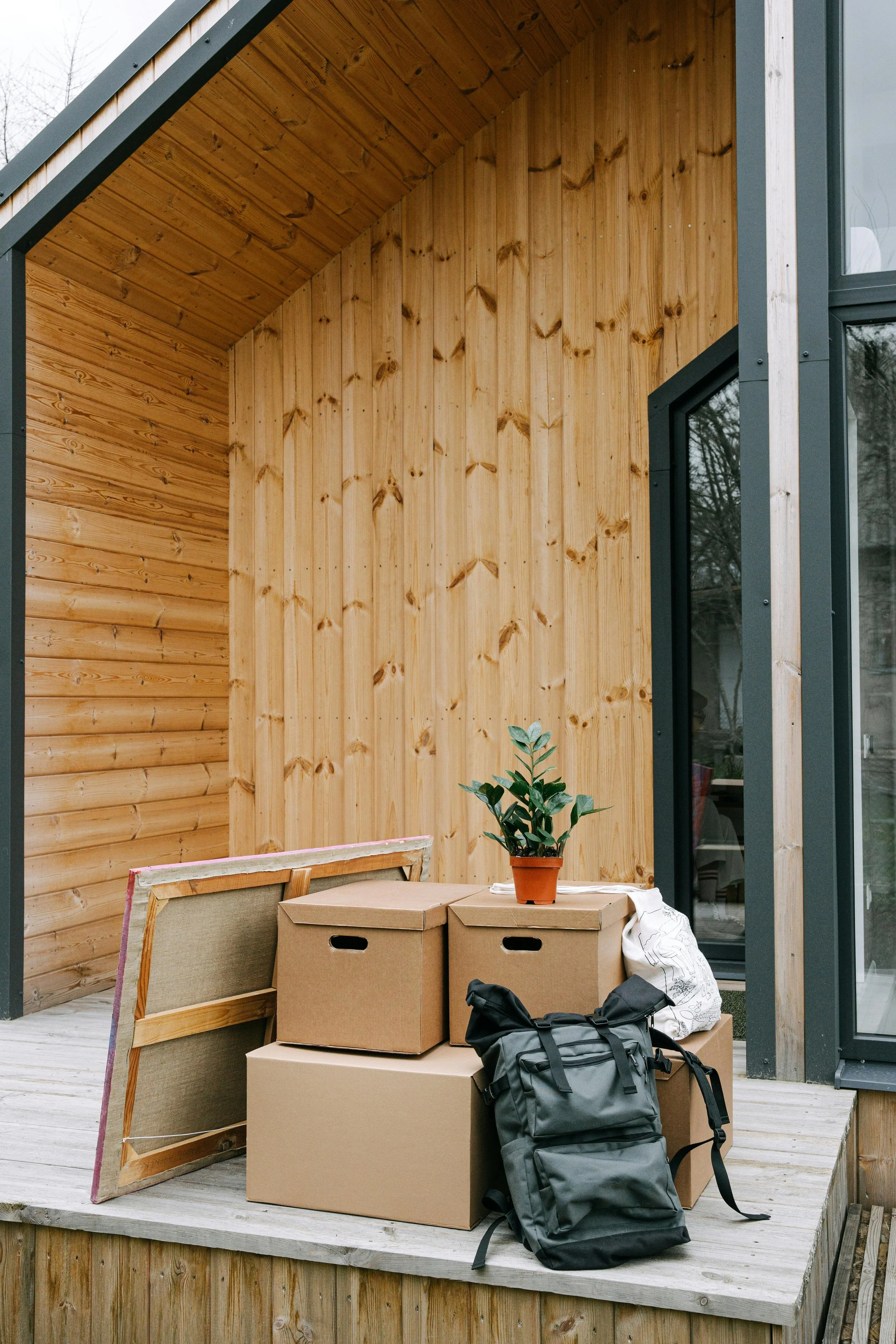 Balcony with wooden walls and ceiling, various cardboard boxes, a potted plant, a black backpack, and a framed mirror leaning against the boxes.