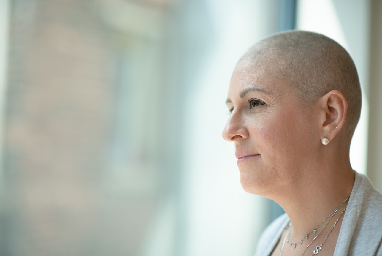 Side profile of a woman with a shaved head wearing pearl earrings, looking out a window with a calm expression.