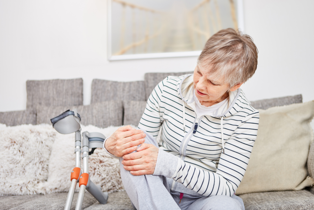 Older woman sitting on a couch, holding her knee in pain, with crutches leaning next to her.