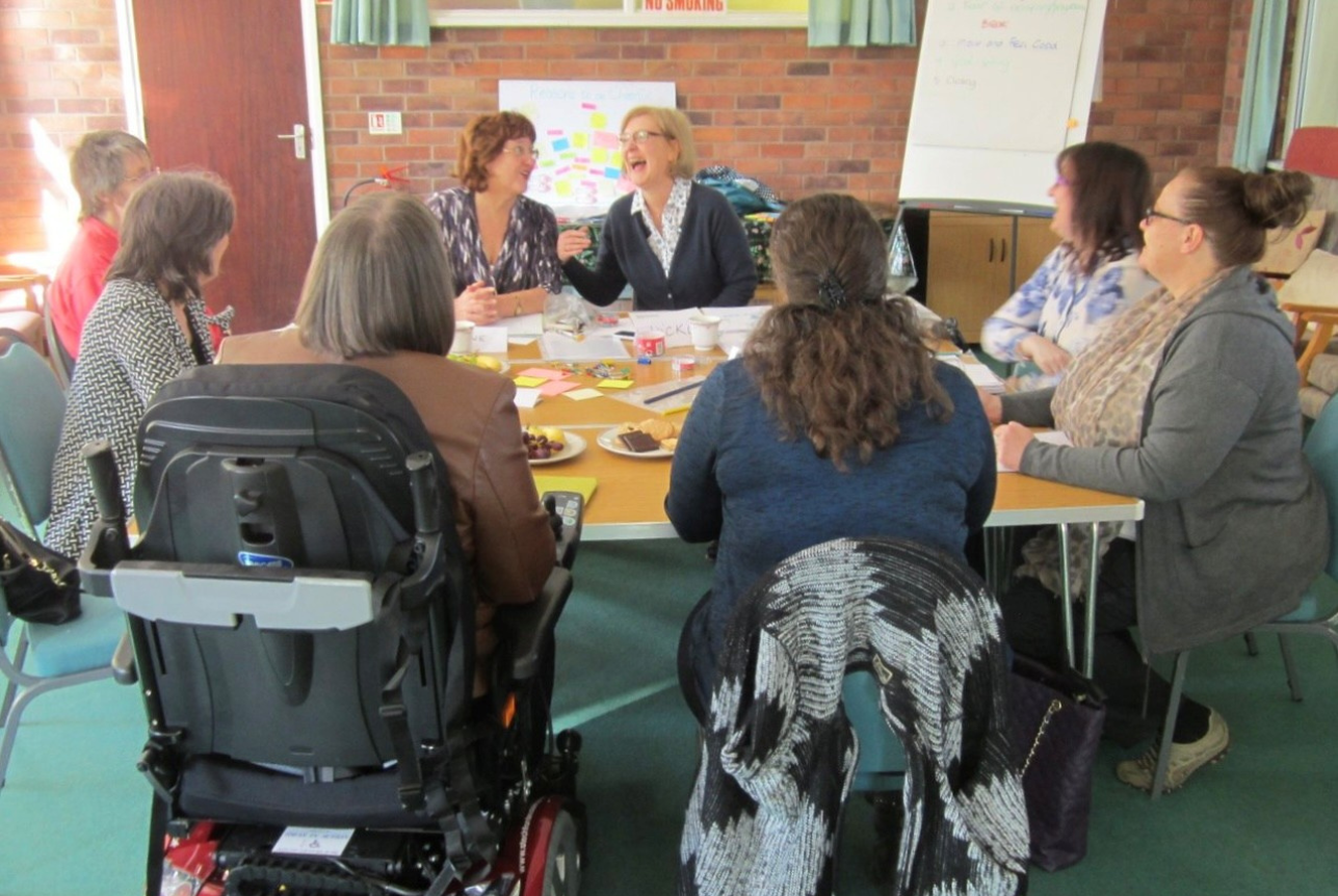 Group of women gathered around a table in a meeting room, laughing and engaging in conversation, with snacks and papers on the table.