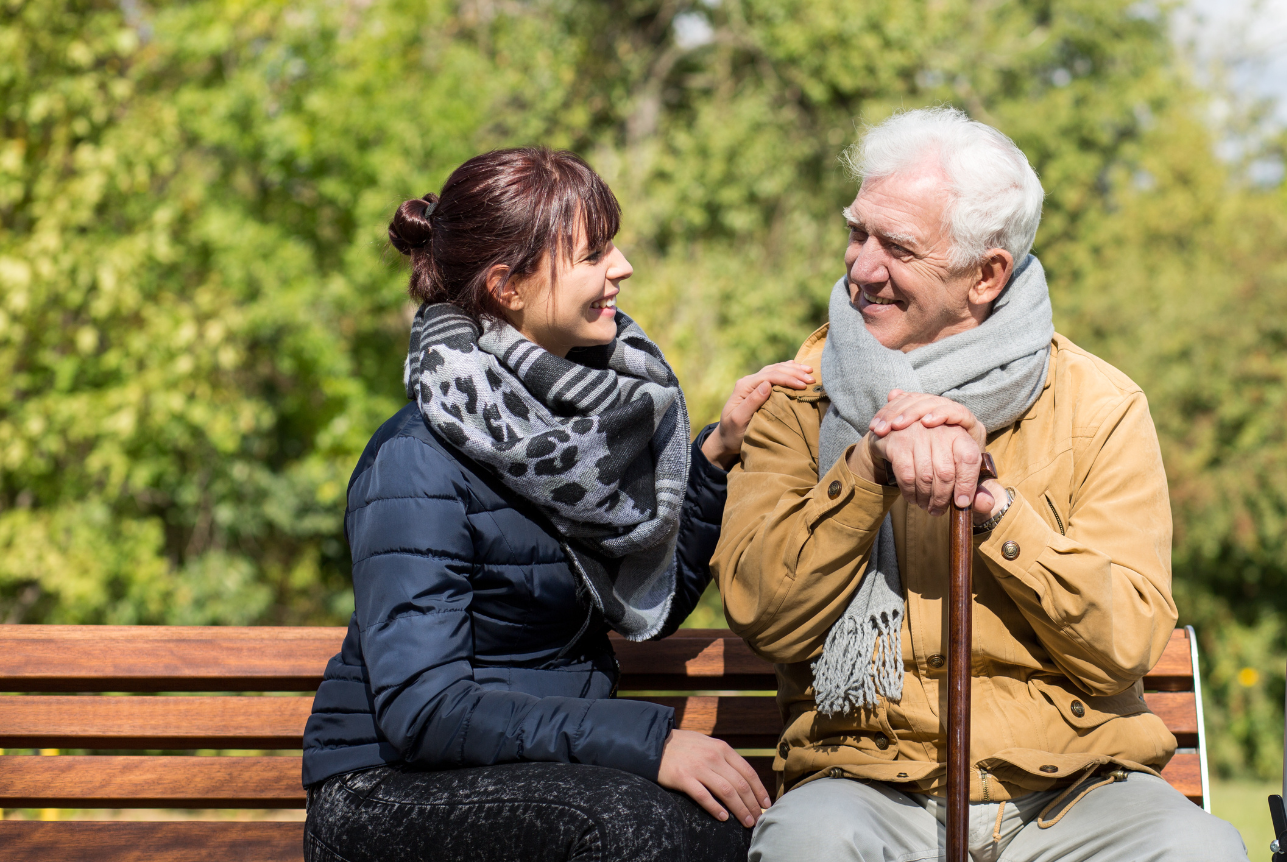 A young woman and an elderly man sitting on a park bench, smiling at each other during daytime.