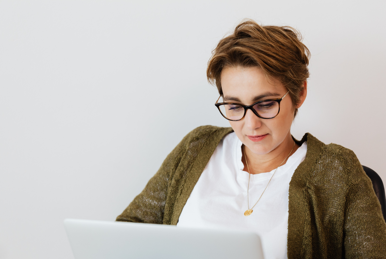 Woman with short brown hair and glasses looking at a laptop screen, wearing a white shirt and brown cardigan, against a plain white wall.