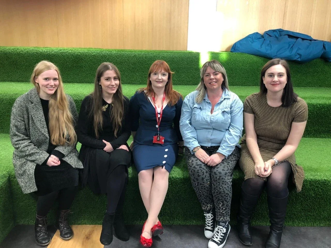 Five women sitting on green artificial grass steps indoors, smiling at the camera.