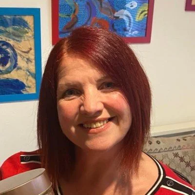 A woman with shoulder-length red hair smiling at the camera, standing in front of colorful artwork on the wall.