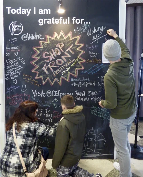 People drawing and writing on a large black chalkboard wall about an upcoming shopfront festival in March.