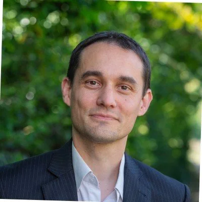 Portrait of a man in a suit outdoors with green trees in the background.