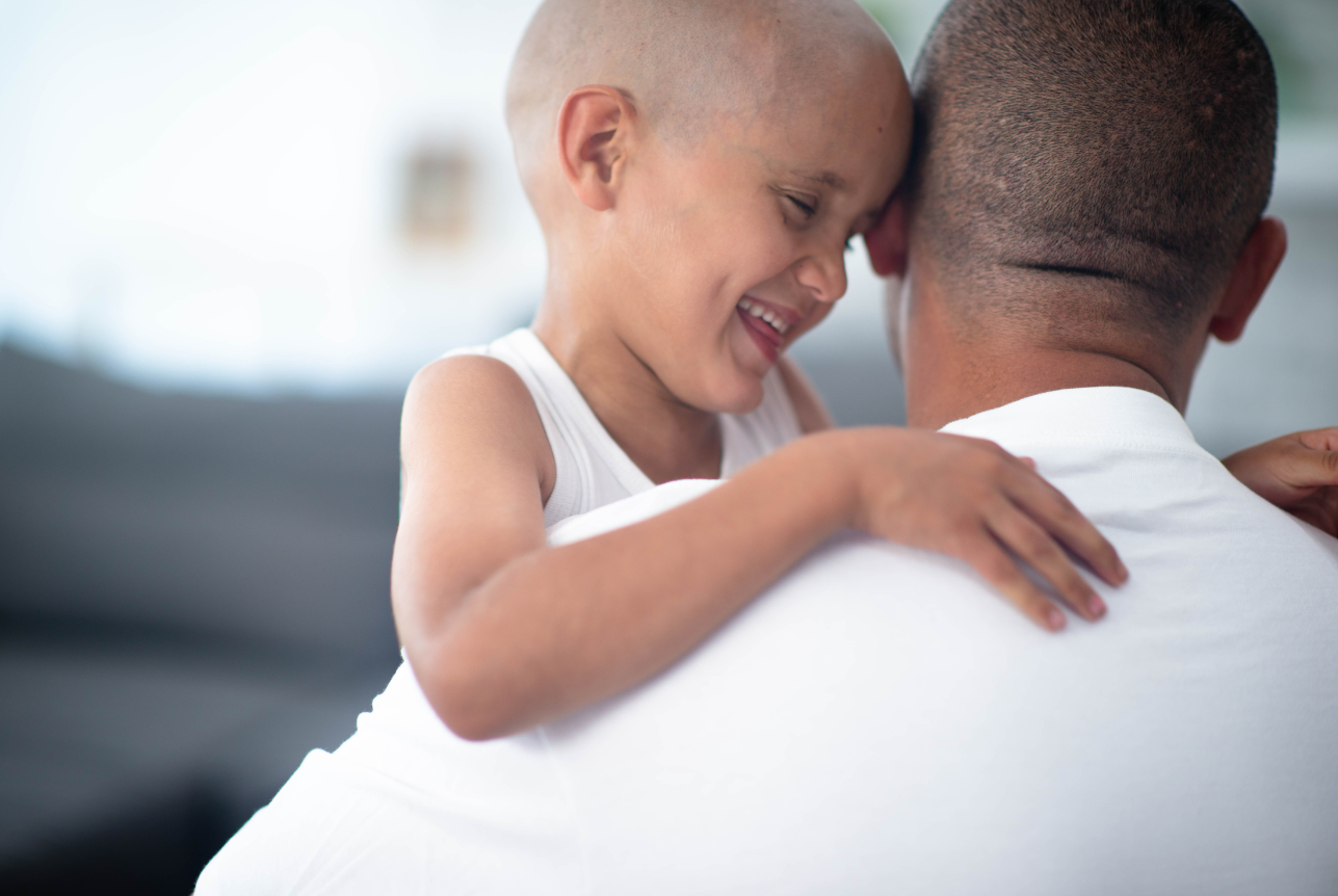 Young girl with a shaved head smiling joyfully while hugging and leaning her forehead on an adult man's shoulder, indoors with soft natural light.