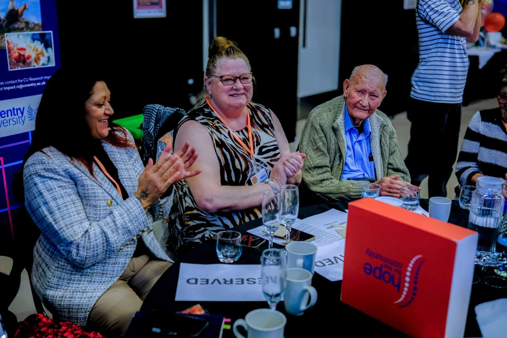 Group of people sitting at a table during a meeting or conference, smiling and clapping, with documents, cups, and glasses on the table.