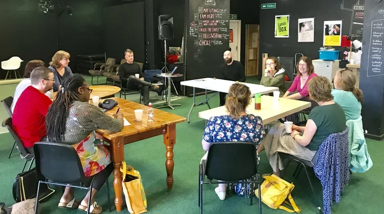 A group of people seated around tables in a room, having a discussion or meeting, with blackboards and posters on the walls.