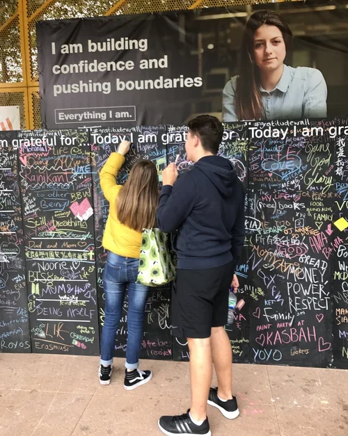 Two teenagers, a girl in a yellow jacket and a boy in a navy hoodie, write messages on a black wall covered with colorful chalk writings and drawings. A large billboard above features a woman with brown hair and a blue shirt, with the text 'I am buil