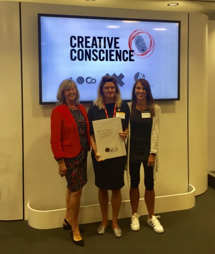 Three women standing in front of a screen that says "Creative Conscience" at a conference or event, with one woman holding a certificate.
