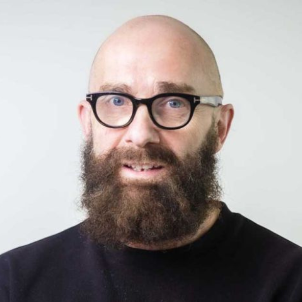 A man with glasses, a bald head, and a full beard, wearing a black shirt, looking at the camera against a plain light background.