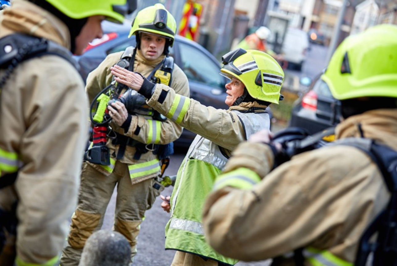 Firefighters in safety gear, including helmets and jackets, engaged in a discussion on a city street, with emergency vehicles and bystanders in the background.