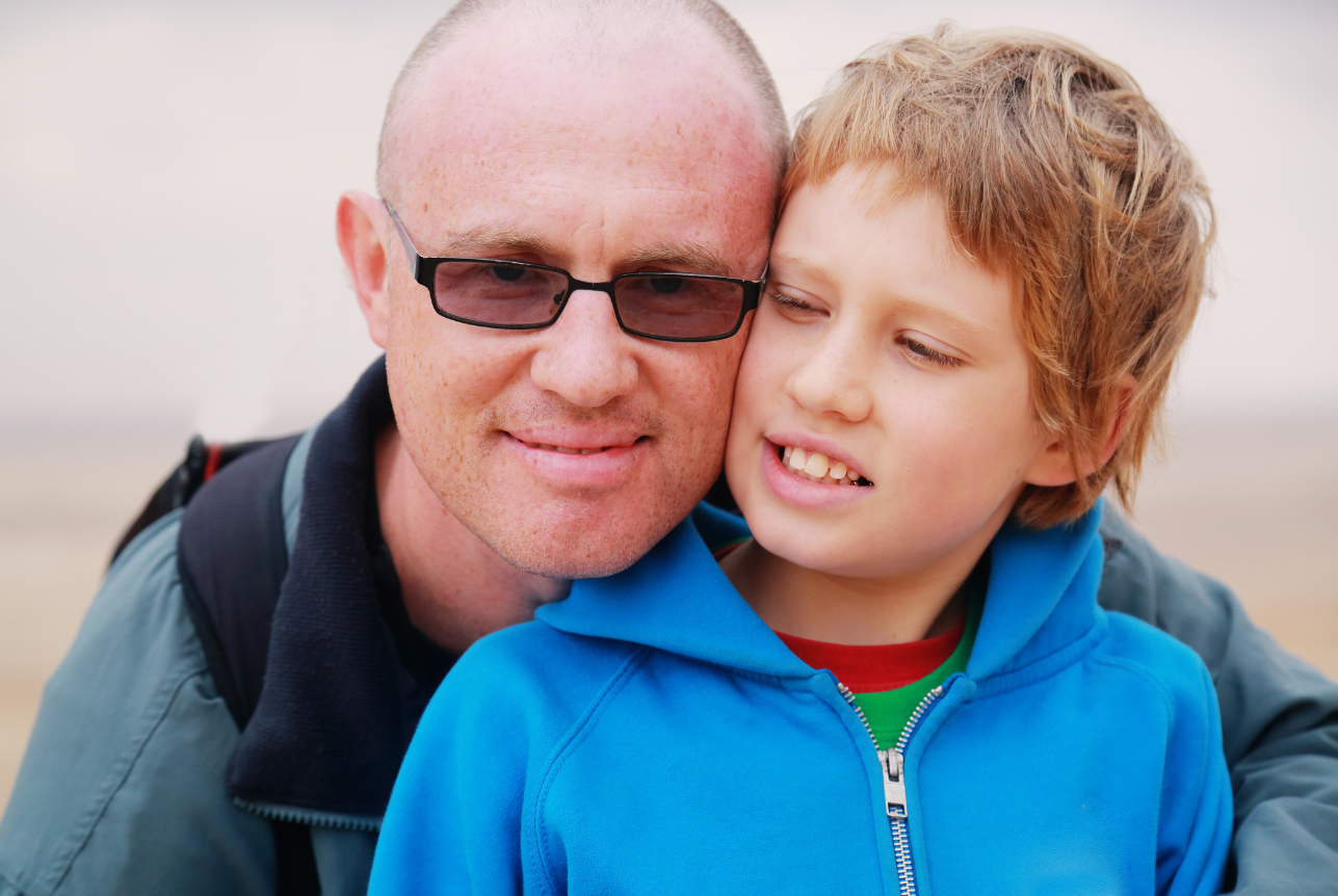 A man with short hair, wearing glasses and a gray jacket, smiling as he leans close to a young boy with light brown hair wearing a bright blue hoodie.