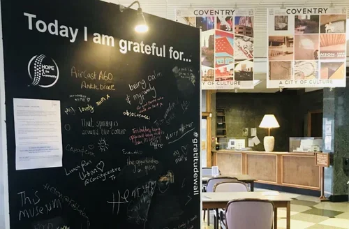 A black chalkboard with the heading 'Today I am grateful for...' filled with handwritten notes. Behind it, there is a wooden counter with a lamp, stacks of papers, and two framed posters labeled 'COVENTRY' on the wall.