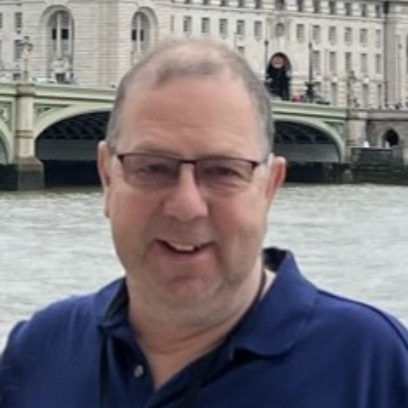 A middle-aged man wearing glasses and a navy blue shirt smiling at the camera, with a river and historic bridge in the background.