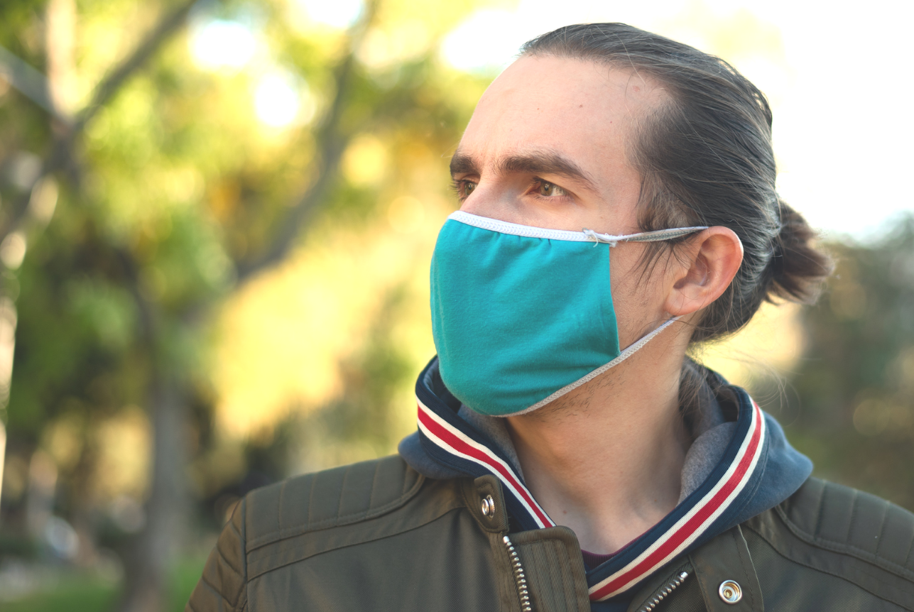 A man with dark hair tied back wearing a teal face mask outdoors, with blurred green and yellow trees in the background.