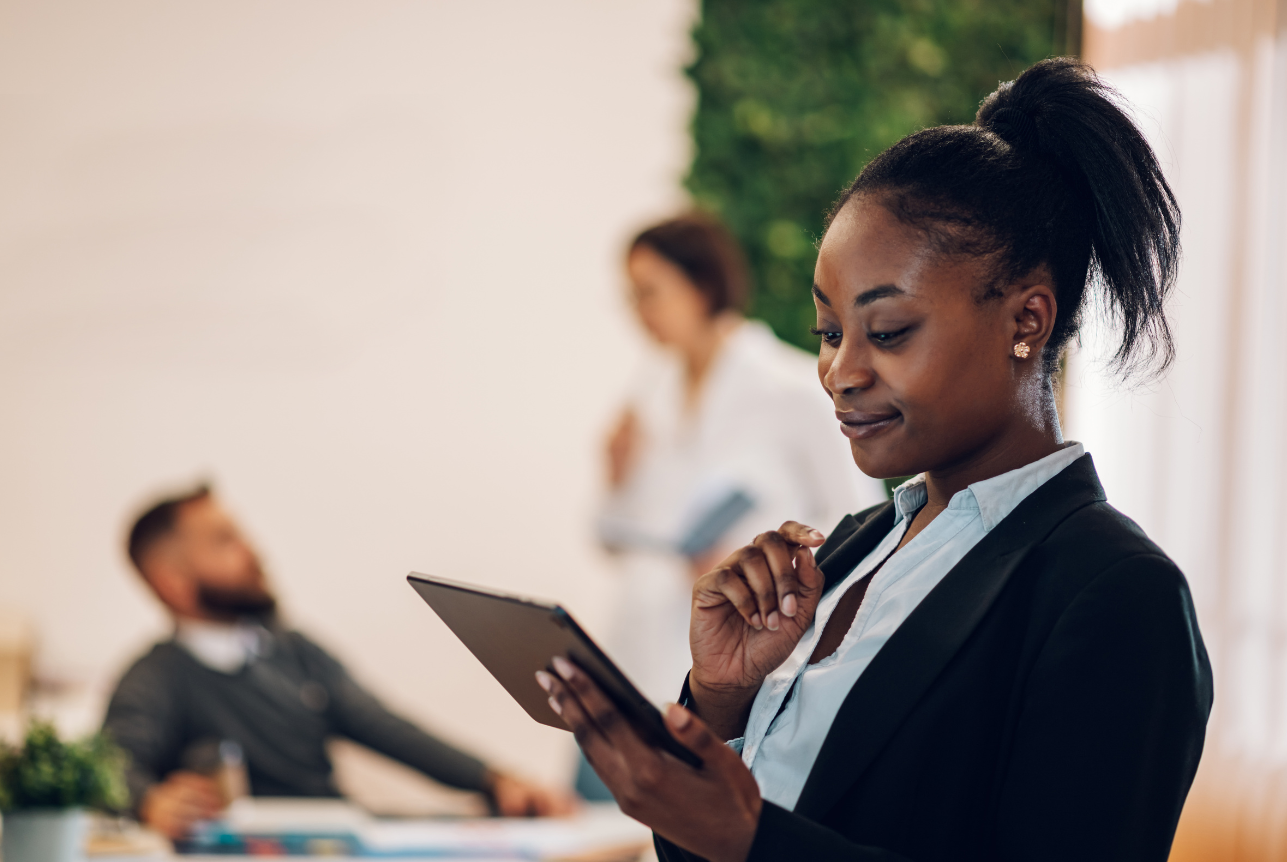Businesswoman in a suit looking at her tablet in an office with colleagues in the background.