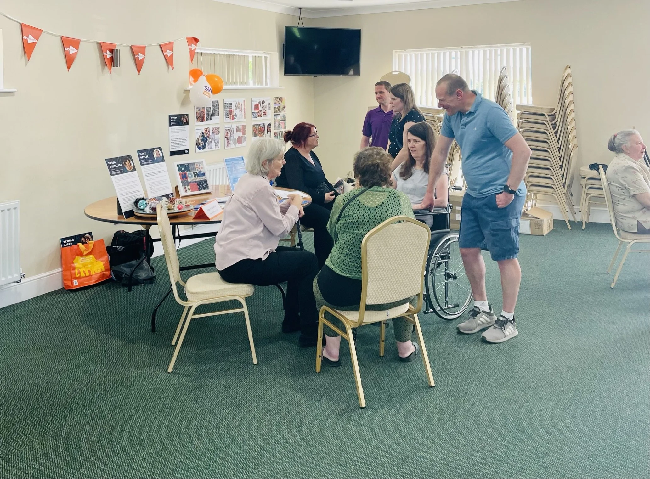 Group of people gathered in a room with decorations and informational displays, some seated and others standing, engaging in conversation.