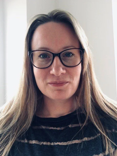 A woman with long, light brown hair, wearing black glasses and a black and gray striped shirt, smiling slightly at the camera against a plain background.