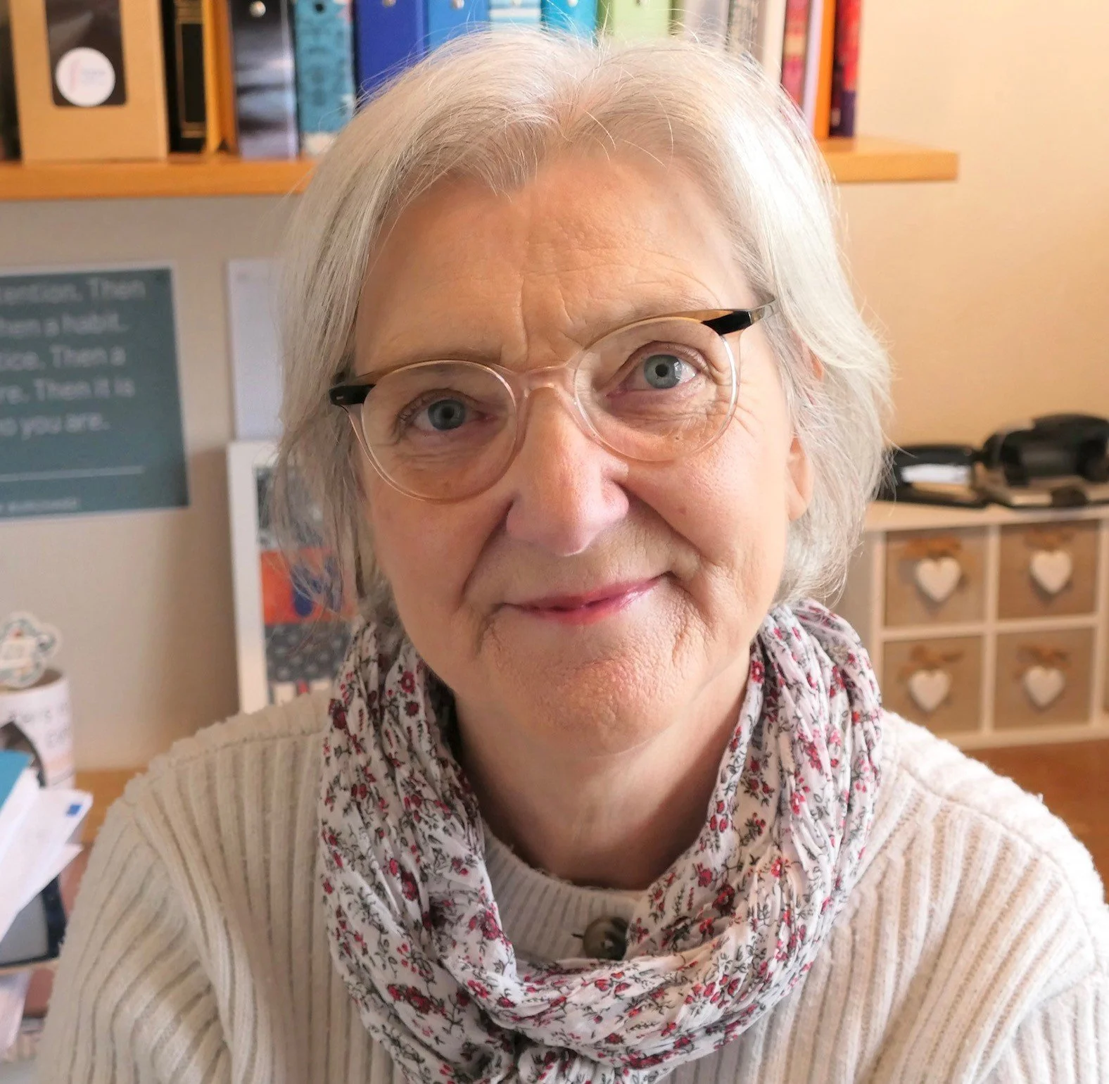 A close-up of an older woman with glasses and light gray hair, smiling slightly, in a room with shelves and a notice board in the background.